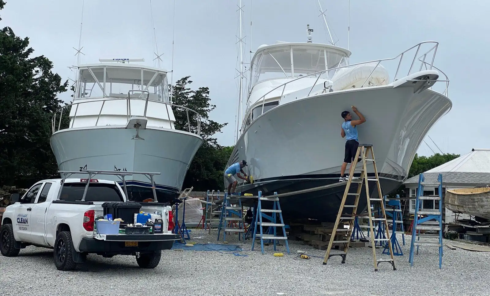Two yachts on stands in a boatyard, one detailer on a ladder polishing a bow, the crew's white pickup parked in the gravel