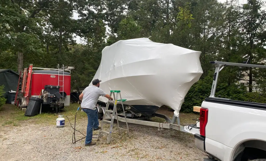 Crew member on a stepladder heat-shrinking white film over a boat on its trailer